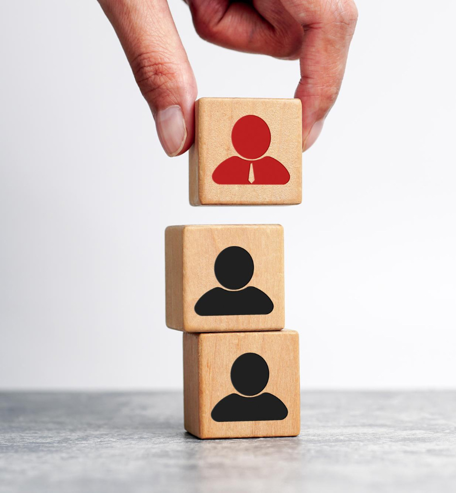 Ręka układające drewniane kostki, dwie na dole mają rysunek czarnej ikony człowieka a ostatnia na górze ma rysunek ikony człowieka ale czerwony i ma krawat