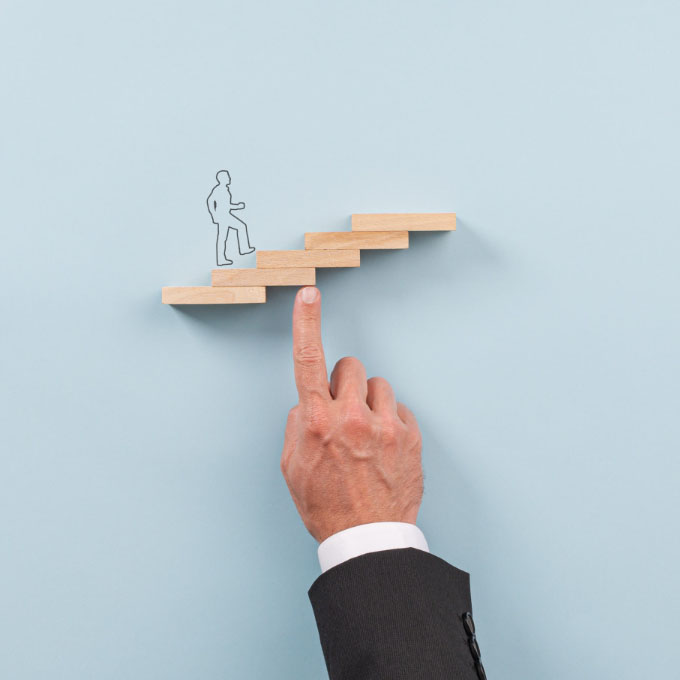 Ręka, której palec podtrzymuje małe drewniane schody, po których wchodzi narysowany człowiek